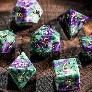 Może przedstawiać: Zestaw siedmiu wielościennych kości do gry w zielono-fioletowym kolorze ze złotymi numerami, umieszczonych w drewnianej misce. Te kości do gier fabularnych obejmują różne kształty, takie jak d20, d12, d10, d8, d6 i d4.