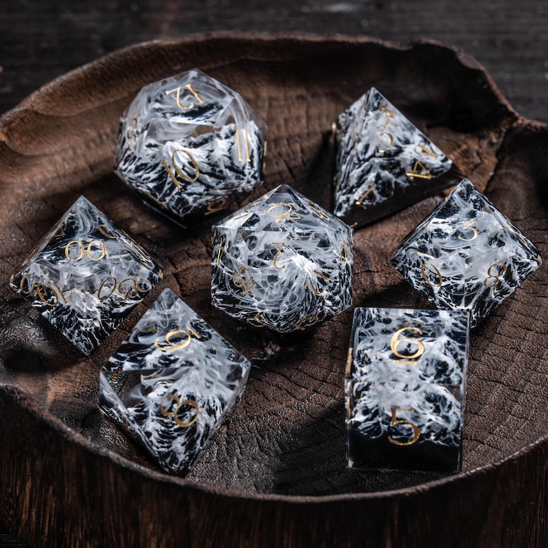 May include: A set of seven polyhedral dice, featuring a black and white marbled pattern with gold numerals. The dice are presented in a wooden bowl. Suitable for role-playing games and other tabletop activities.