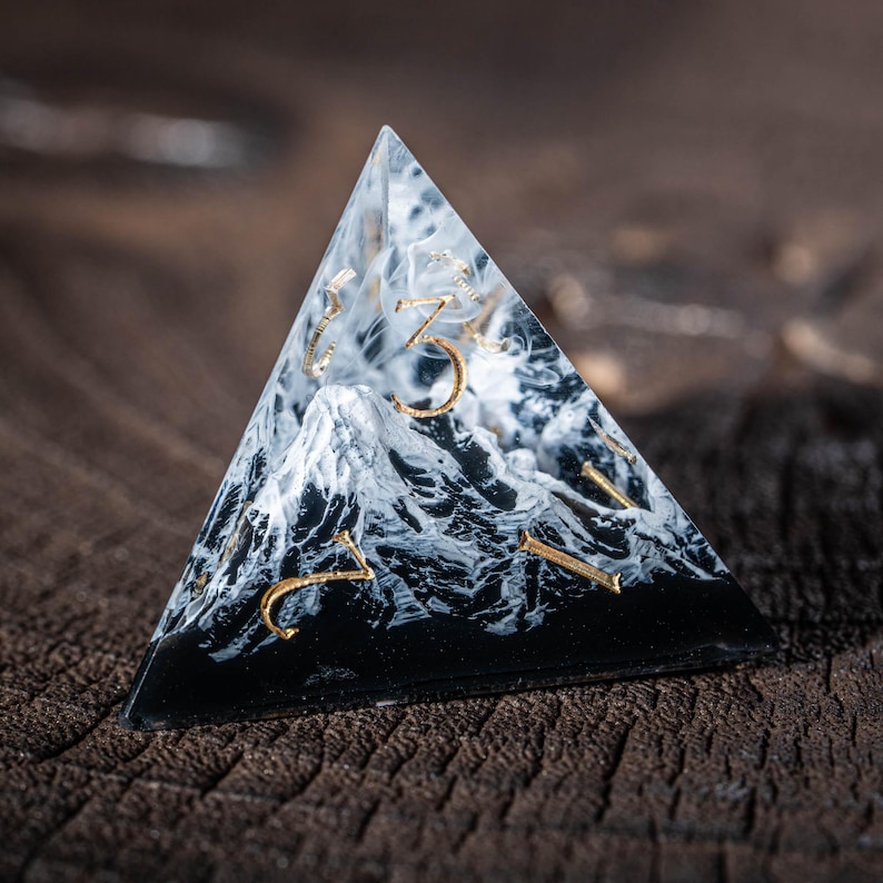 May include: A pyramid-shaped die made of clear resin, featuring a black and white marbled design inside, reminiscent of mountains. Gold numerals are visible on the faces. The die rests on a dark wooden surface.