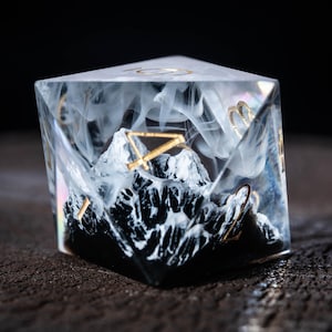 May include: A clear, multi-sided die featuring a miniature mountain range. The mountains are black and white, with gold detailing and numbers. White, smoky material fills the die, creating a fantasy theme. The die is set against a dark background.