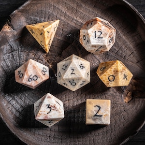 May include: A collection of seven multi-sided dice, each with a unique shape and number of faces. The dice are made of a stone-like material with a marbled beige and brown pattern. The numbers are in black. The dice are arranged on a dark wooden surface.