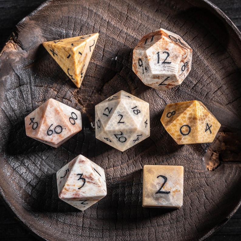 May include: A collection of seven multi-sided dice, each with a unique shape and number of faces. The dice are made of a stone-like material with a marbled beige and brown pattern. The numbers are in black. The dice are arranged on a dark wooden surface.
