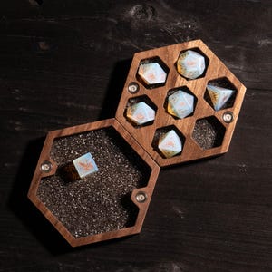 Puede incluir: Una caja de madera con tapa hexagonal, que contiene siete dados iridiscentes con detalles dorados. Los dados est&aacute;n dispuestos en forma de panal dentro de la caja.