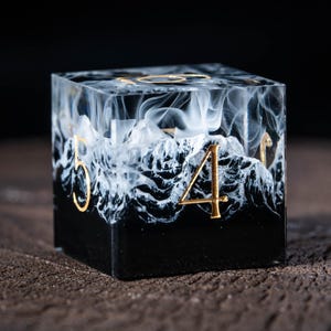 May include: A six-sided die with a black base and a white, cloud-like design. Gold numerals are visible on the sides. The die is made of a clear resin material.