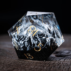 May include: A clear, multi-sided die with a mountain range design. The interior features a black and white mountain scene with gold-coloured numbers. The die is designed for tabletop gaming.
