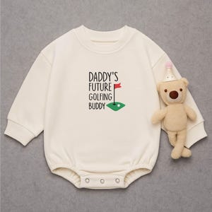 May include: Cream-colored baby onesie with the text "DADDY'S FUTURE GOLFING BUDDY" and a golf-themed graphic. A small teddy bear wearing a party hat is next to the onesie. The onesie has long sleeves and snap closures.
