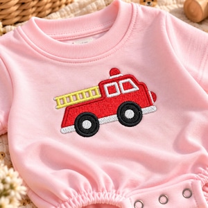 Puede incluir: Un body de manga corta en color rosa claro con un bordado de camión de bomberos rojo en el pecho. El camión tiene una escalera amarilla, ventanas blancas y ruedas negras. La prenda presenta un cuello redondo acanalado y tres broches metálicos.