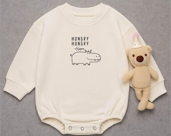 May include: Cream-colored baby onesie with long sleeves and snap closures. The onesie features a black line drawing of a hippo and the words "HUNGRY HUNGRY Hippo". A small teddy bear wearing a party hat is next to the onesie.