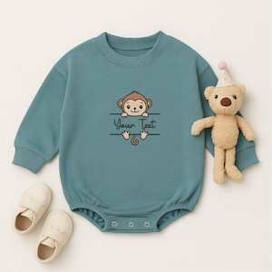 May include: A teal baby romper with a crew neck and long sleeves. The romper features a cartoon monkey graphic with the text "Your Text". A small stuffed bear with a party hat and a pair of baby shoes are next to the romper.