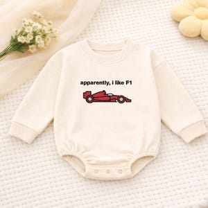 Könnte beinhalten: Cremefarbener Baby-Strampler mit langen Ärmeln. Der Strampler zeigt den Text "apparently, I like F1" über einer roten Rennwagen-Grafik. Das Kleidungsstück hat Druckknöpfe am unteren Ende.