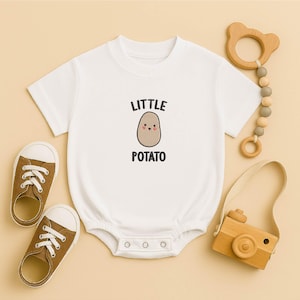 以下が含まれることがあります： 「LITTLE POTATO」の文字とジャガイモのイラストが入った白いベビーボディスーツ。茶色のキャンバススニーカー、木製のカメラのおもちゃ、木製のクマ型歯固めも写っています。