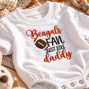 Könnte beinhalten: Ein weißer Kurzarm-Body mit einer orangefarbenen und schwarzen Stickerei mit der Aufschrift Bengals FAN JUST LIKE daddy. Eine braun-weiße Grafik eines Footballs befindet sich zwischen den Wörtern Bengals und FAN auf der Vorderseite.