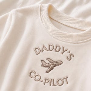 May include: A cream-coloured crewneck sweatshirt featuring brown embroidered text that reads DADDY'S CO-PILOT, with an embroidered aeroplane graphic positioned between the two lines of text, all centred on the chest of the garment.