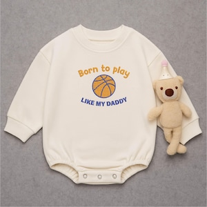 Può includere: Pagliaccetto color crema per neonati con maniche lunghe e chiusure a scatto. Il pagliaccetto presenta la scritta ricamata "Born to play LIKE MY DADDY" sopra una grafica di basket. Un piccolo orsacchiotto con un cappello da festa è accanto al pagliaccetto.