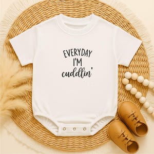 May include: White baby onesie with the text "EVERYDAY I'M cuddlin'" in black script. The onesie is paired with brown leather baby shoes and a beaded necklace, all on a woven mat.