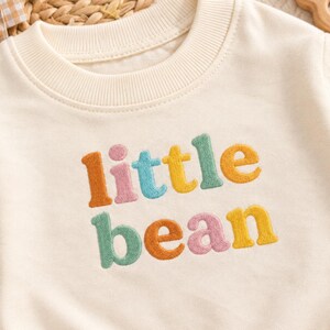 May include: A cream-coloured crewneck sweatshirt featuring the embroidered text "little bean" across the chest. The letters are rendered in a puffy, textured style using a palette of muted orange, pink, teal, and yellow thread colours.
