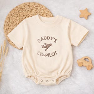 Könnte beinhalten: Cremefarbener Baby-Strampler mit kurzen Ärmeln und rundem Halsausschnitt. Der Strampler hat den gestickten Text "DADDY'S CO-PILOT" mit einer Flugzeug-Grafik. Holzstern und Spielzeugauto im Hintergrund.
