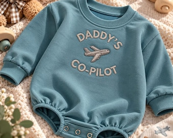 Puede incluir: Un body de bebé de manga larga en color azul polvoriento con cuello redondo acanalado y puños. El pecho muestra el texto bordado DADDY'S CO-PILOT con un pequeño gráfico de avión en el centro. La parte inferior incluye tres broches metálicos.