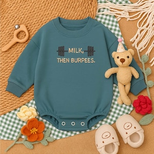 May include: Teal baby onesie with the text "MILK, THEN BURPEES" in a barbell design. The long-sleeved onesie has a round neckline and snap closures. A teddy bear wearing a party hat and baby slippers are next to the onesie.