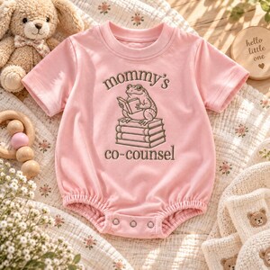 May include: A light pink short-sleeved baby bodysuit featuring an embroidered graphic of a frog sitting on a stack of books. The text "mommy's co-counsel" is embroidered in a dark brown thread above and below the frog illustration.