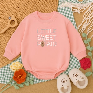 May include: A light pink baby bodysuit with the text "LITTLE SWEET POTATO" embroidered on the front. The long-sleeved one-piece has snap closures at the bottom. The image also includes a pair of white baby shoes.