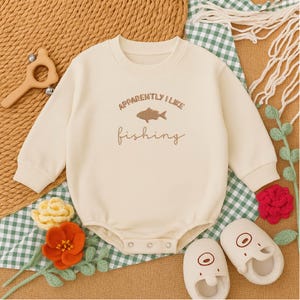 May include: Cream-colored baby bodysuit with long sleeves and the text "Apparently I Like Fishing" in brown. The bodysuit features a fish graphic. The image includes matching baby shoes and decorative elements.