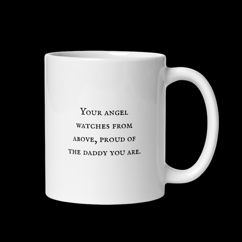 Puede incluir: Una taza de cer&aacute;mica blanca con asa curva. La taza presenta el texto negro "Your angel watches from above, proud of the daddy you are" centrado en la parte delantera. La taza est&aacute; sobre un fondo negro.