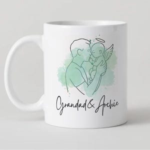 Puede incluir: Taza de cer&aacute;mica blanca con un dibujo lineal de una persona sosteniendo a un beb&eacute; con alas de &aacute;ngel y un halo. La taza tiene el texto "Grandad & Archie" en una fuente manuscrita, con un lavado de acuarela verde en el fondo.