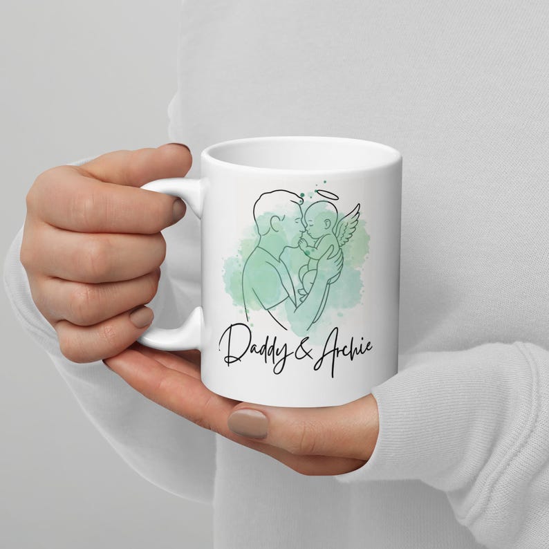 Puede incluir: Taza de cer&aacute;mica blanca con una ilustraci&oacute;n lineal de un padre sosteniendo a un beb&eacute; con alas de &aacute;ngel y un halo, sobre un fondo de acuarela turquesa. La taza tiene el texto "Daddy & Archie" en una fuente cursiva.