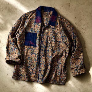以下が含まれることがあります： ゴールド、レッド、ブルーの色合いで構成された、密度の高いマルチカラーの花柄が特徴の長袖ボタンダウンシャツ。無地のダークブルーの襟と、小さな赤い刺繍が施された同色の胸ポケットが付いています。
