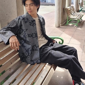 May include: A long-sleeved button-down shirt featuring a dark gray and black geometric pattern, worn over a plain cream-colored t-shirt paired with black trousers. The shirt has a standard collar and a buttoned front closure with long sleeves.