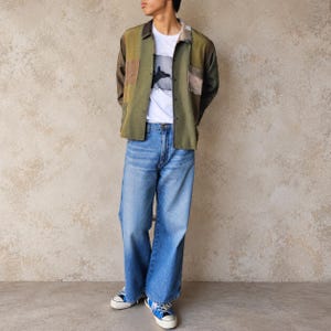 May include: A long-sleeve button-down shirt in olive green with brown patch pockets, worn over a white t-shirt with a dark graphic print. The outfit includes loose-fit blue denim jeans and blue and white low-top canvas sneakers with white laces.