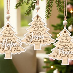 May include: Three wooden Christmas tree decorations shaped like trees, with a light wood grain and white trim. Each has a star and the text "Unto us a child is born...the Prince of Peace". They hang from natural twine with a wooden bead.