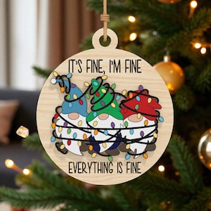 May include: Wooden Christmas ornament featuring three gnomes with colorful hats and string lights. The ornament has the text "IT'S FINE, I'M FINE, EVERYTHING IS FINE" in black lettering. The ornament is hanging on a Christmas tree.
