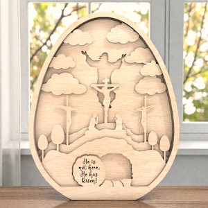 Puede incluir: Decoración de madera en forma de huevo de Pascua con un diseño en capas que representa la crucifixión, la resurrección y el texto "He is not here, He has Risen!". La obra de arte presenta nubes, árboles y figuras, todo en un tono de madera natural.