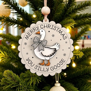 May include: A round, light-colored Christmas ornament with a scalloped edge, featuring a cartoon goose wearing a bow tie. The text "MERRY CHRISTMAS YOU SILLY GOOSE" is printed around the goose. The ornament hangs on a Christmas tree.