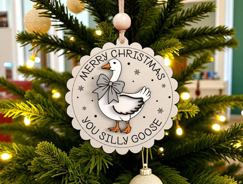 May include: A round, light-colored Christmas ornament with a scalloped edge, featuring a cartoon goose wearing a bow tie. The text "MERRY CHRISTMAS YOU SILLY GOOSE" is printed around the goose. The ornament hangs on a Christmas tree.