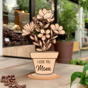 Pode incluir: Um arranjo floral de madeira em um vaso com as palavras "I Love You Mom" gravadas na frente. As flores são cortadas a laser com detalhes intrincados. O arranjo fica em uma base de madeira, perfeito para uma mesa ou prateleira.