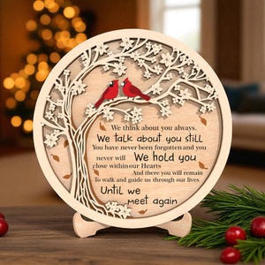 May include: A wooden memorial plaque with a tree design, two red cardinals, and the text "We think about you always... Until we meet again." The plaque is round and stands on a wooden base, with a blurred background.