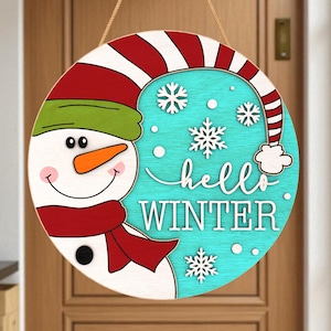 May include: Circular wooden snowman sign with a red scarf, green hat, and orange nose. The sign features the words "hello WINTER" in white script on a turquoise background, with white snowflakes. A festive winter decoration.