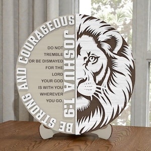 May include: A round wooden decorative piece with a lion's face on one half and inspirational text on the other. The text reads "Be Strong and Courageous" and a Bible verse. The piece is supported by a wooden stand.