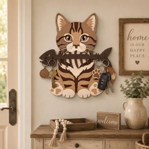 May include: A wooden cat key holder in shades of brown and tan, featuring a striped pattern. The cat holds a key rack with keychains, including a yarn ball, paw print, and keys. A small wooden sign that says "welcome" is on the shelf below.