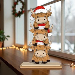 Peut inclure: Décoration de Noël en bois représentant trois vaches des Highlands empilées, chacune portant un bonnet de Père Noël. Les vaches sont marron avec des visages et des cornes clairs. La décoration repose sur une base en bois.