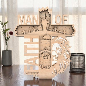 Puede incluir: Arte de pared de madera en forma de cruz con las palabras "MAN OF FAITH" recortadas. El diseño incluye un león, un pesebre y el versículo bíblico "2 Corintios 5:7". El arte está hecho de madera de color claro.