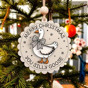 May include: A round, light gray Christmas ornament with a scalloped edge. The ornament features a cartoon goose wearing a gray bow tie, with the text "MERRY CHRISTMAS YOU SILLY GOOSE" in a circular pattern. The ornament is hanging on a Christmas tree.