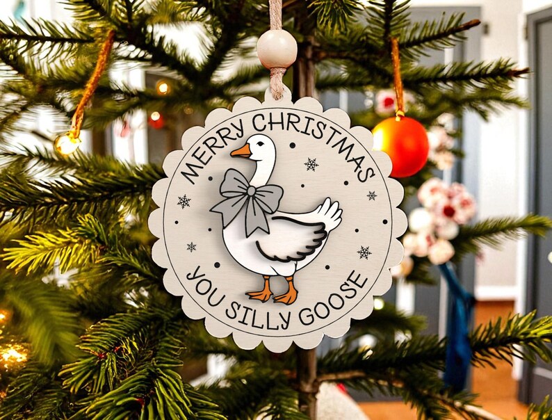 May include: A round, light gray Christmas ornament with a scalloped edge. The ornament features a cartoon goose wearing a gray bow tie, with the text "MERRY CHRISTMAS YOU SILLY GOOSE" in a circular pattern. The ornament is hanging on a Christmas tree.