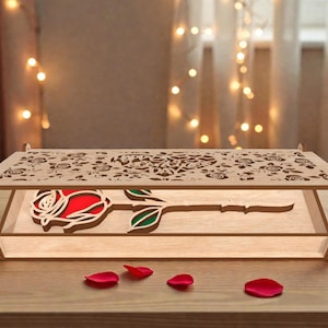 Könnte beinhalten: Eine Holzkiste mit einem lasergeschnittenen Rosenmuster. Die Kiste hat einen Klappdeckel mit floralen Ausschnitten und eine rote Rosen-Silhouette im Inneren. Die Kiste ist aus hellem Holz gefertigt und steht auf einer Holzoberfläche. Rote Rosenblätter sind verstreut.