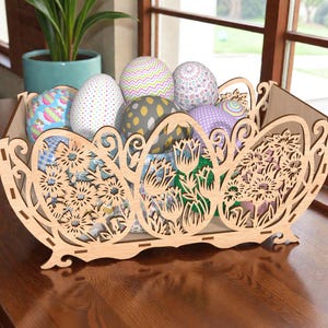 May include: A wooden Easter basket with laser-cut floral designs, filled with patterned Easter eggs. The basket is made of light-colored wood and sits on a dark wooden surface. The eggs have various patterns and colors.