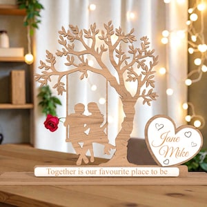 May include: A wooden decorative piece featuring a tree with a swing holding two figures. A heart-shaped element with the names "Jane Mike" and the phrase "Together is our favourite place to be" are also included. A red rose is placed on the left.