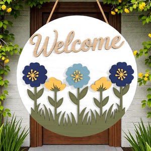 Puede incluir: Un letrero de bienvenida redondo de madera con fondo blanco. La palabra "Welcome" está escrita en letras marrones claras. Debajo, recortes de flores azules y amarillas con tallos y hierba verdes. El letrero cuelga de una cuerda marrón.